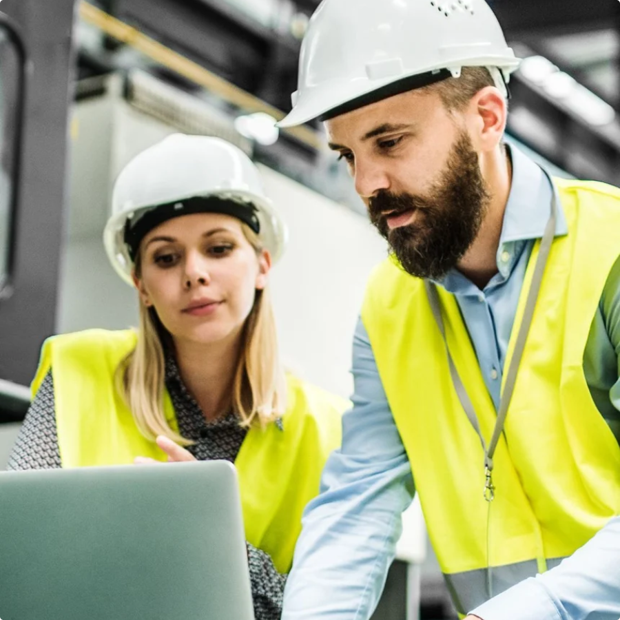 Two industrial workers in hard hats and safety vests reviewing data on a laptop in a manufacturing facility. Two industrial workers in hard hats and safety vests reviewing data on a laptop in a manufacturing facility.